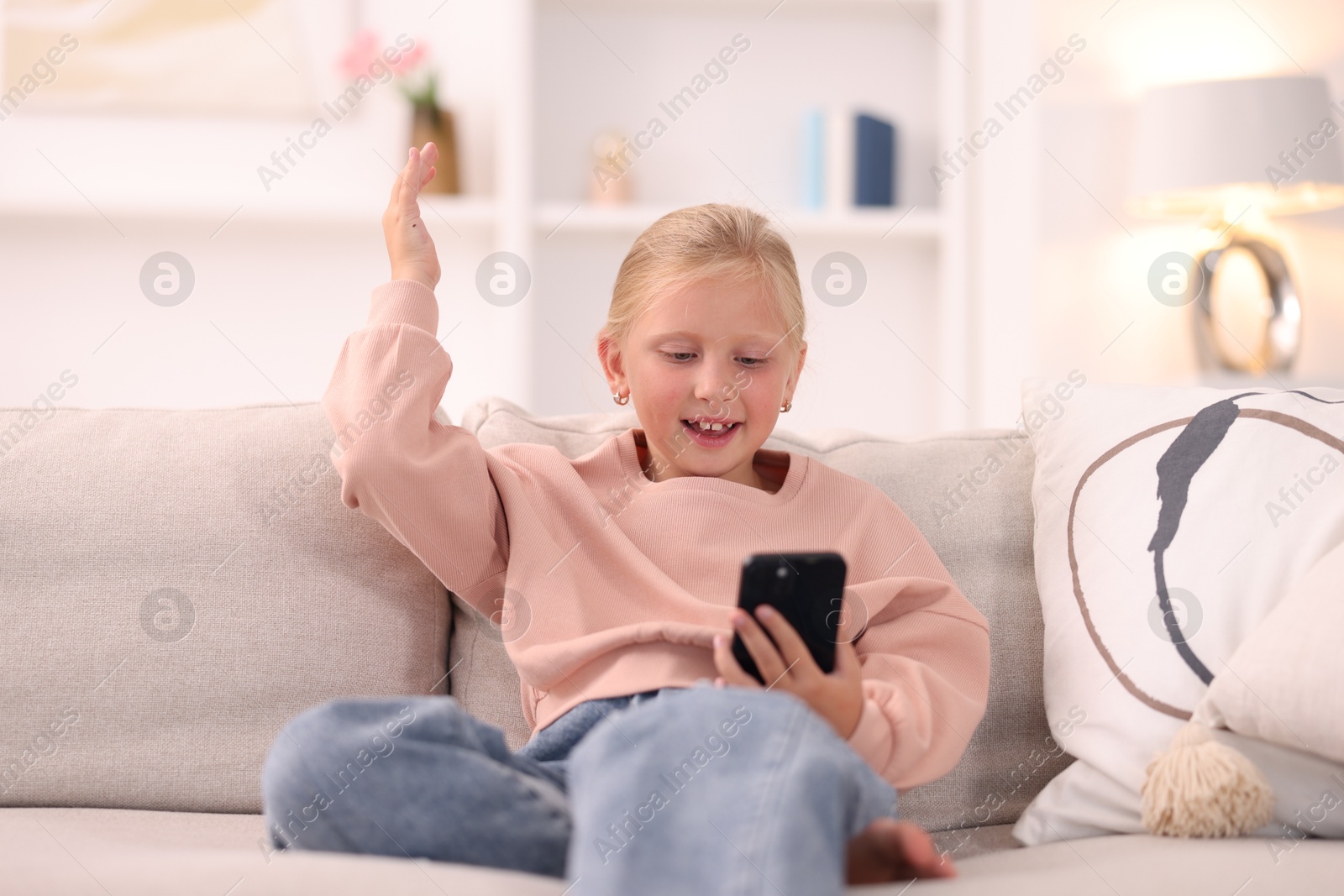 Photo of Cute little girl with smartphone laughing on sofa at home