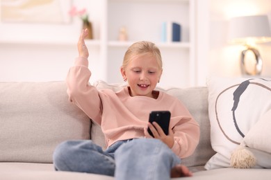 Photo of Cute little girl with smartphone laughing on sofa at home
