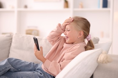 Photo of Cute little girl with smartphone laughing on sofa at home