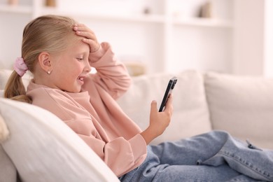 Photo of Cute little girl with smartphone laughing on sofa at home