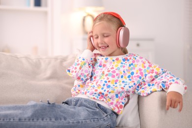 Photo of Smiling little girl in headphones listening to music on sofa at home