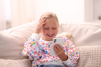 Photo of Cute little girl with smartphone laughing on sofa at home