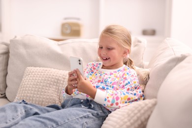 Photo of Cute little girl with smartphone laughing on sofa at home