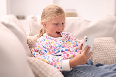 Photo of Emotional little girl with smartphone on sofa at home