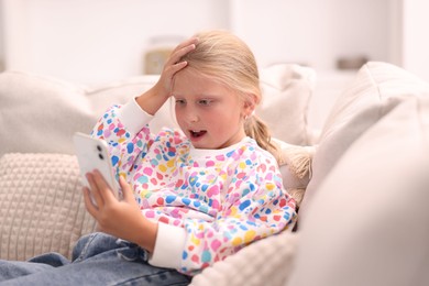 Photo of Emotional little girl with smartphone on sofa at home