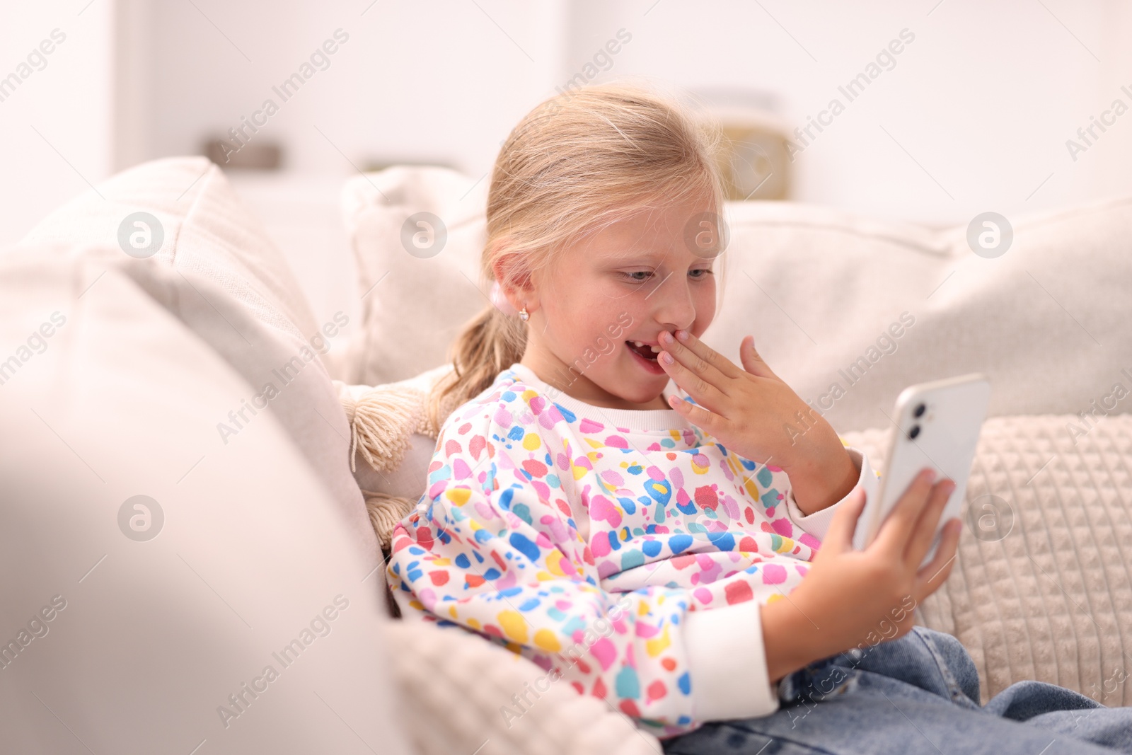 Photo of Cute little girl with smartphone covering mouth and laughing on sofa at home