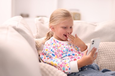 Photo of Cute little girl with smartphone covering mouth and laughing on sofa at home