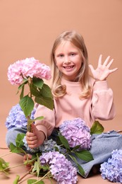 Photo of Smiling little girl with hydrangea flowers waving hello on brown background