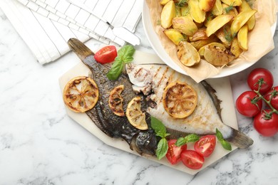 Photo of Tasty roasted flounder fish served with potatoes, tomatoes, basil and lemon slices on white marble table, flat lay. Homemade seafood dish