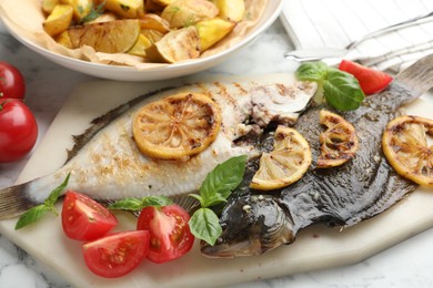 Photo of Tasty roasted flounder fish served with potatoes, tomatoes, basil and lemon slices on white marble table, closeup. Homemade seafood dish