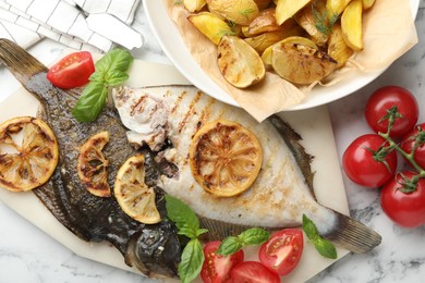 Photo of Tasty roasted flounder fish served with potatoes, tomatoes, basil and lemon slices on white marble table, flat lay. Homemade seafood dish