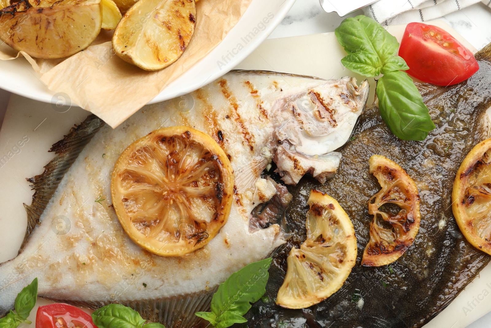 Photo of Tasty roasted flounder fish served with potatoes, tomatoes, basil and lemon slices on white marble table, flat lay. Homemade seafood dish