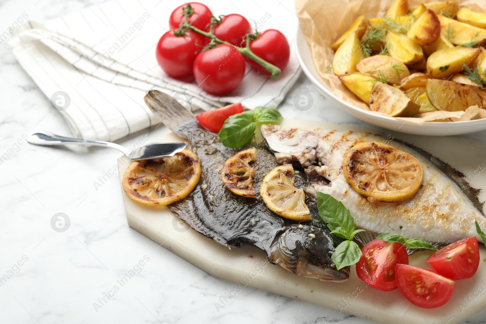 Photo of Tasty roasted flounder fish served with potatoes, tomatoes, basil and lemon slices on white marble table, closeup. Homemade seafood dish