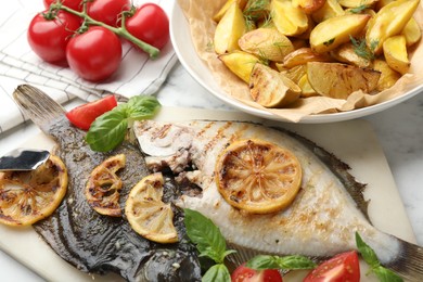 Photo of Tasty roasted flounder fish served with potatoes, tomatoes, basil and lemon slices on white marble table, closeup. Homemade seafood dish
