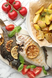 Photo of Tasty roasted flounder fish served with potatoes, tomatoes, basil and lemon slices on white marble table, flat lay. Homemade seafood dish