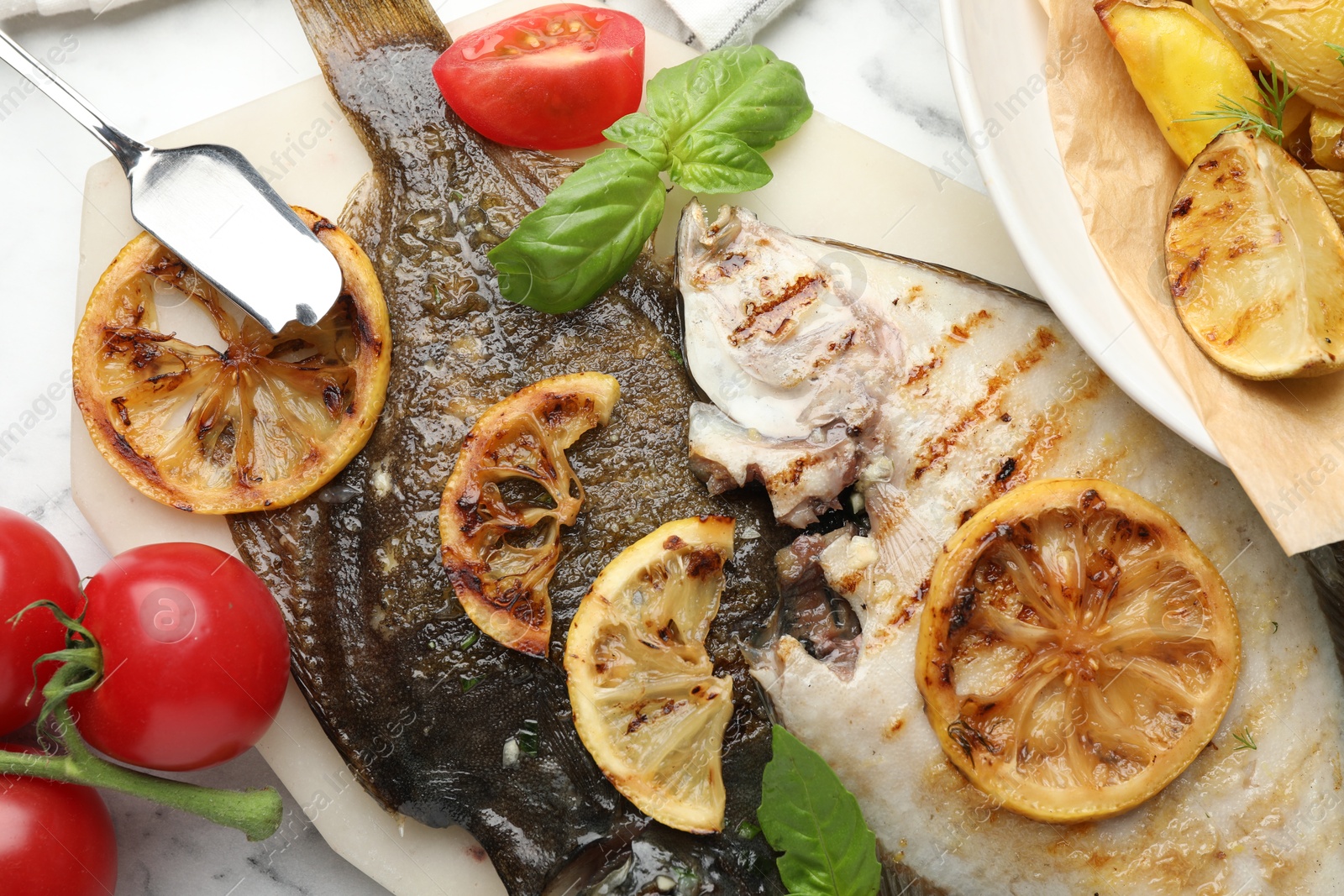 Photo of Tasty roasted flounder fish served with potatoes, tomatoes, basil and lemon slices on white marble table, flat lay. Homemade seafood dish