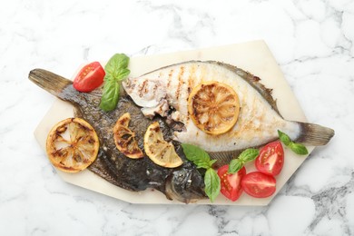 Photo of Tasty roasted flounder fish served with tomatoes, basil and lemon slices on white marble table, top view. Homemade seafood dish