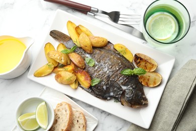 Photo of Tasty roasted flounder fish served with potatoes, basil and lemon slices on white marble table, flat lay. Homemade seafood dish