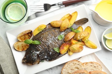 Photo of Tasty roasted flounder fish served with potatoes, basil and lemon slices on white marble table, flat lay. Homemade seafood dish