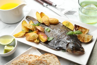 Photo of Tasty roasted flounder fish served with potatoes, basil and lemon slices on white marble table, closeup. Homemade seafood dish