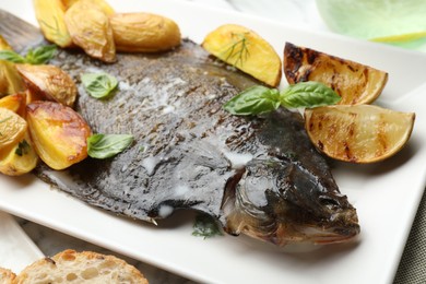 Photo of Tasty roasted flounder fish served with potatoes, basil and lemon slices on white table, closeup. Homemade seafood dish