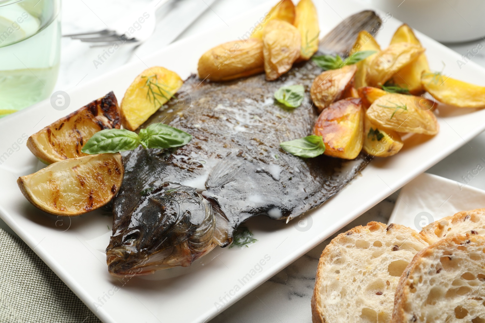 Photo of Tasty roasted flounder fish served with potatoes, basil and lemon slices on white marble table, closeup. Homemade seafood dish