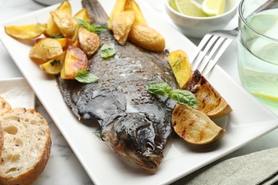 Photo of Tasty roasted flounder fish served with potatoes, basil and lemon slices on white marble table, closeup. Homemade seafood dish