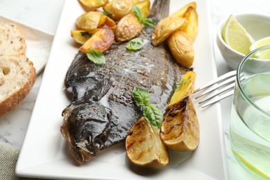 Photo of Tasty roasted flounder fish served with potatoes, basil and lemon slices on white marble table, closeup. Homemade seafood dish