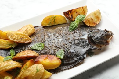 Photo of Tasty roasted flounder fish served with potatoes, basil and lemon slices on white marble table, closeup. Homemade seafood dish