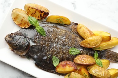 Photo of Tasty roasted flounder fish served with potatoes, basil and lemon slices on white marble table, top view. Homemade seafood dish