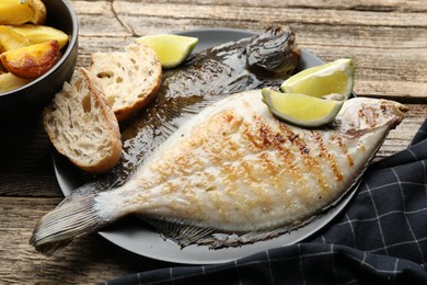 Photo of Tasty roasted flounder fish served with potatoes, bread and lime slices on wooden table, closeup. Homemade seafood dish