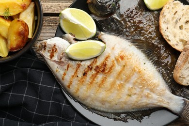 Photo of Tasty roasted flounder fish served with potatoes, bread and lime slices on wooden table, flat lay. Homemade seafood dish