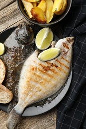 Photo of Tasty roasted flounder fish served with potatoes, bread and lime slices on wooden table, flat lay. Homemade seafood dish