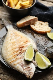 Photo of Tasty roasted flounder fish served with potatoes, bread and lime slices on wooden table, closeup. Homemade seafood dish