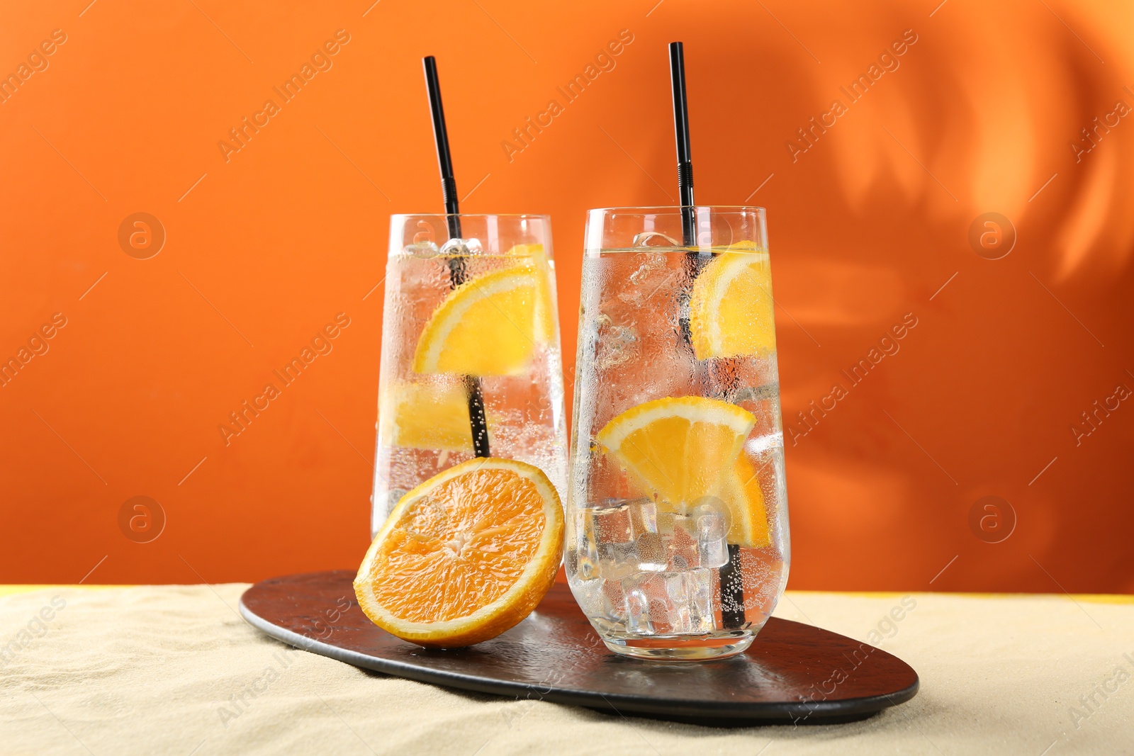 Photo of Refreshing soda water with orange slices and ice cubes in glasses on sand against orange background