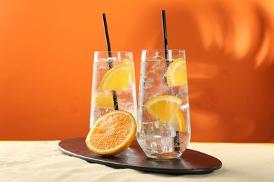 Photo of Refreshing soda water with orange slices and ice cubes in glasses on sand against orange background