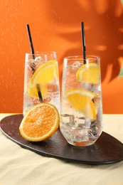 Photo of Refreshing soda water with orange slices and ice cubes in glasses on sand against orange background