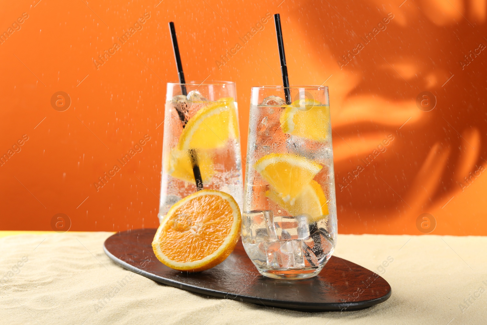 Photo of Refreshing soda water with orange slices and ice cubes in glasses on sand against orange background