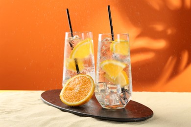 Photo of Refreshing soda water with orange slices and ice cubes in glasses on sand against orange background