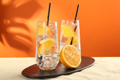 Photo of Refreshing soda water with orange slices and ice cubes in glasses on sand against orange background