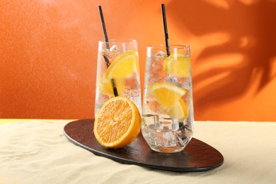 Photo of Refreshing soda water with orange slices and ice cubes in glasses on sand against orange background
