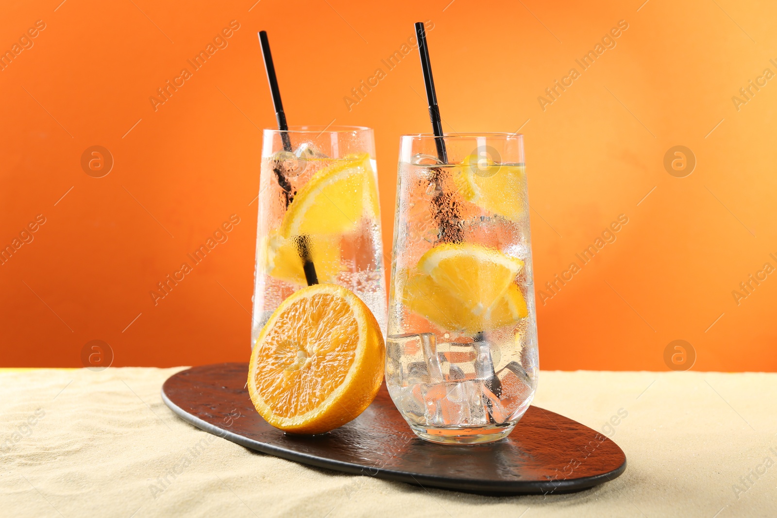 Photo of Refreshing soda water with orange slices and ice cubes in glasses on sand against orange background
