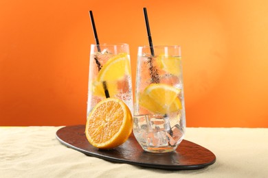 Photo of Refreshing soda water with orange slices and ice cubes in glasses on sand against orange background