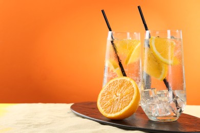 Photo of Refreshing soda water with orange slices and ice cubes in glasses on sand against orange background, closeup. Space for text