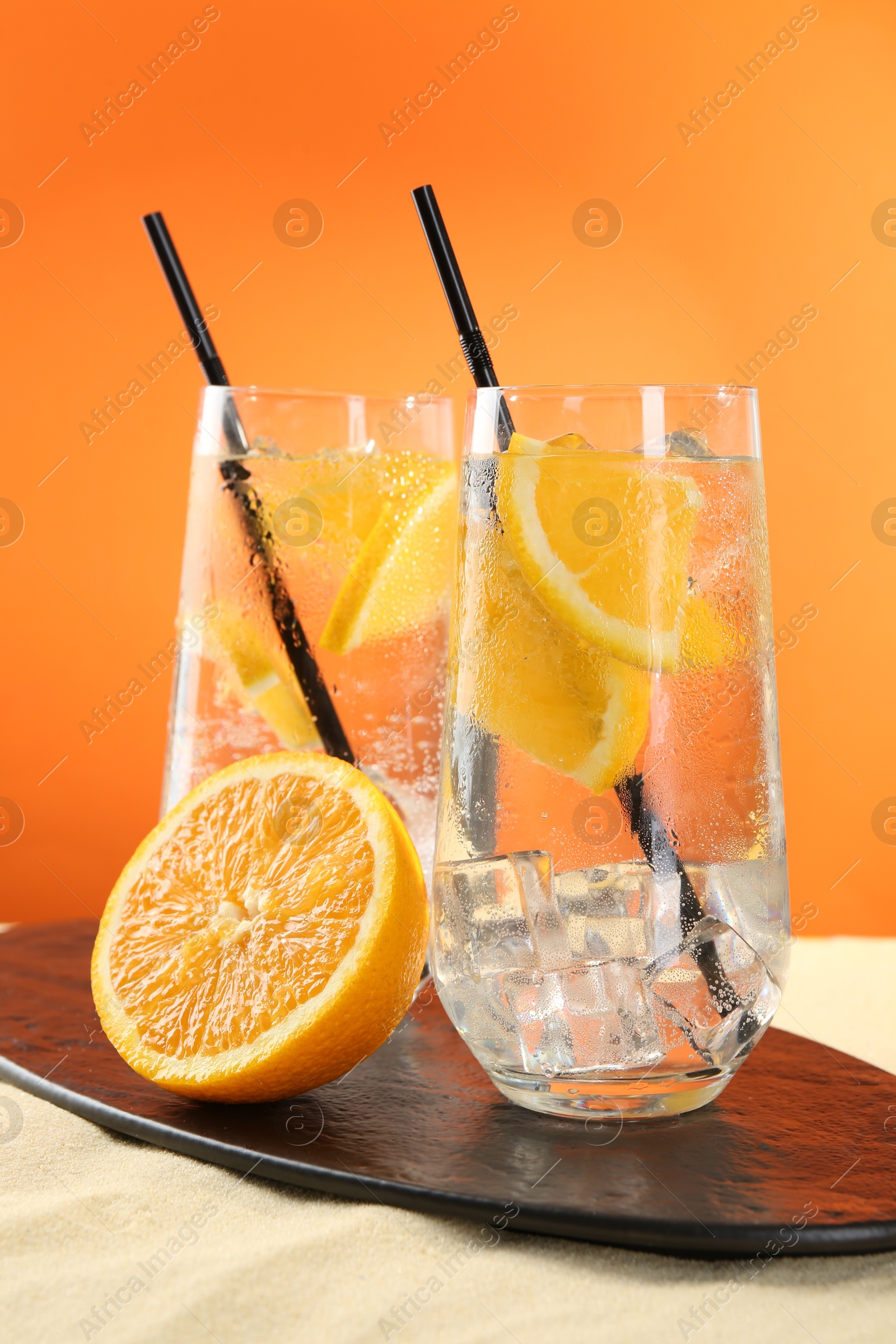 Photo of Refreshing soda water with orange slices and ice cubes in glasses on sand against orange background, closeup