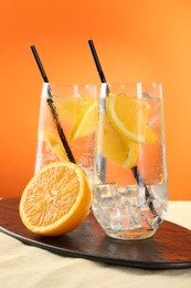 Photo of Refreshing soda water with orange slices and ice cubes in glasses on sand against orange background, closeup