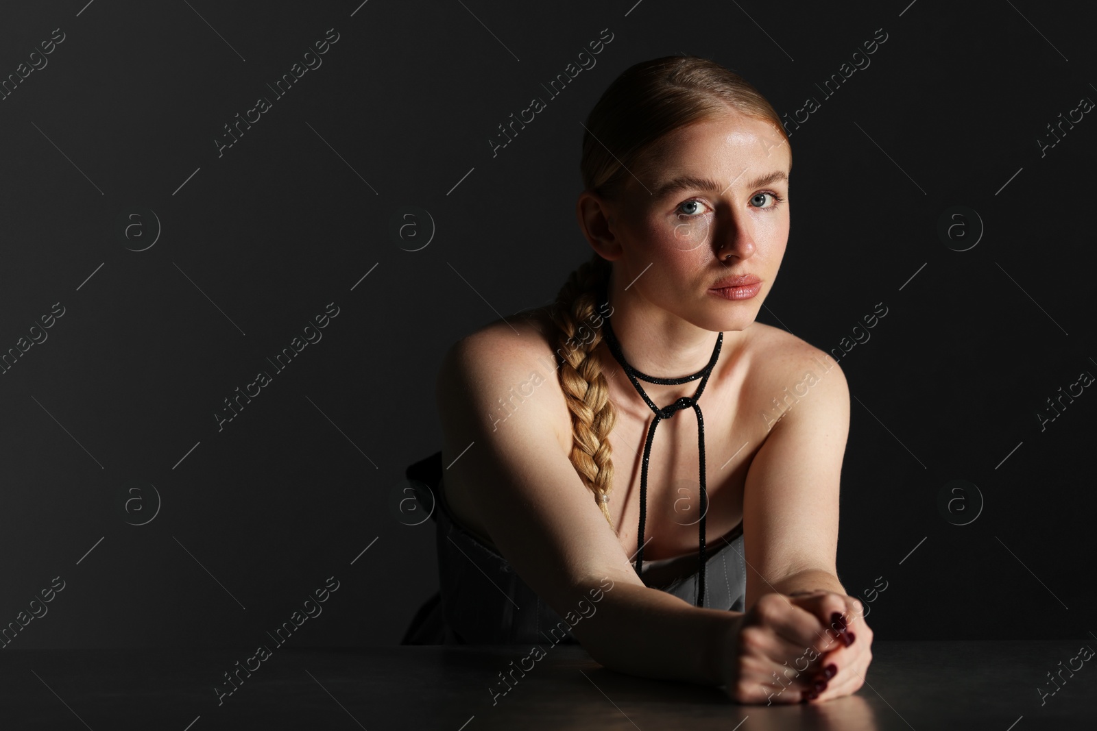 Photo of Fashionable portrait of beautiful woman with pigtail posing at table on dark background. Space for text