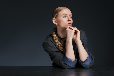 Photo of Beautiful woman with pigtail in suit posing at table on dark background. Space for text