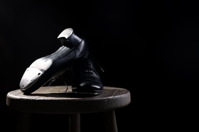 Photo of Pair of shoes for tap dance on wooden stool against black background, closeup. Space for text