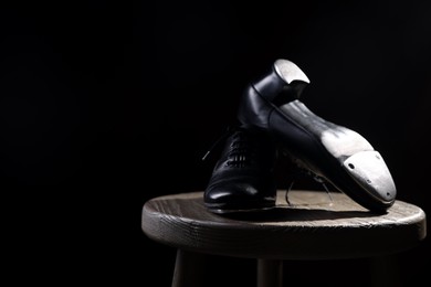 Photo of Pair of shoes for tap dance on wooden stool against black background, closeup. Space for text